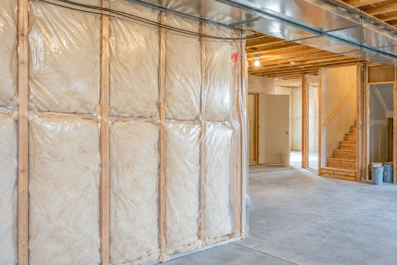 Fiberglass insulation covered with clear plastic fills the exposed wall cavities of an unfinished basement beside a concrete floor and stairs.