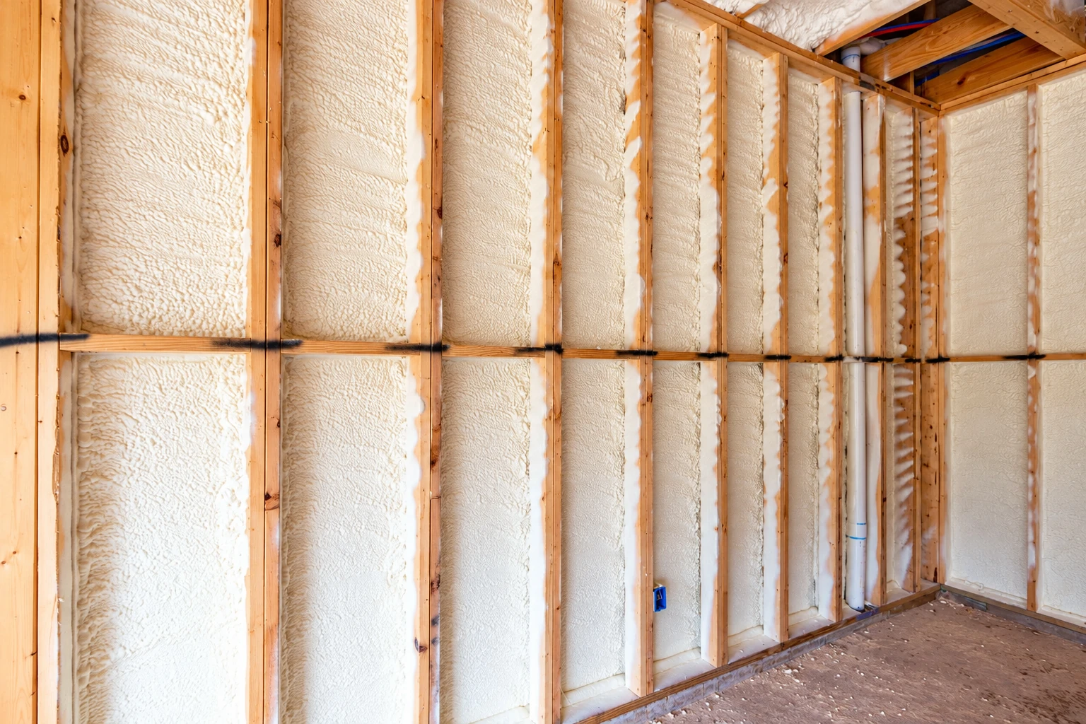 Wall framing filled with cream-colored closed-cell spray foam, with plumbing pipes and electrical boxes visible.