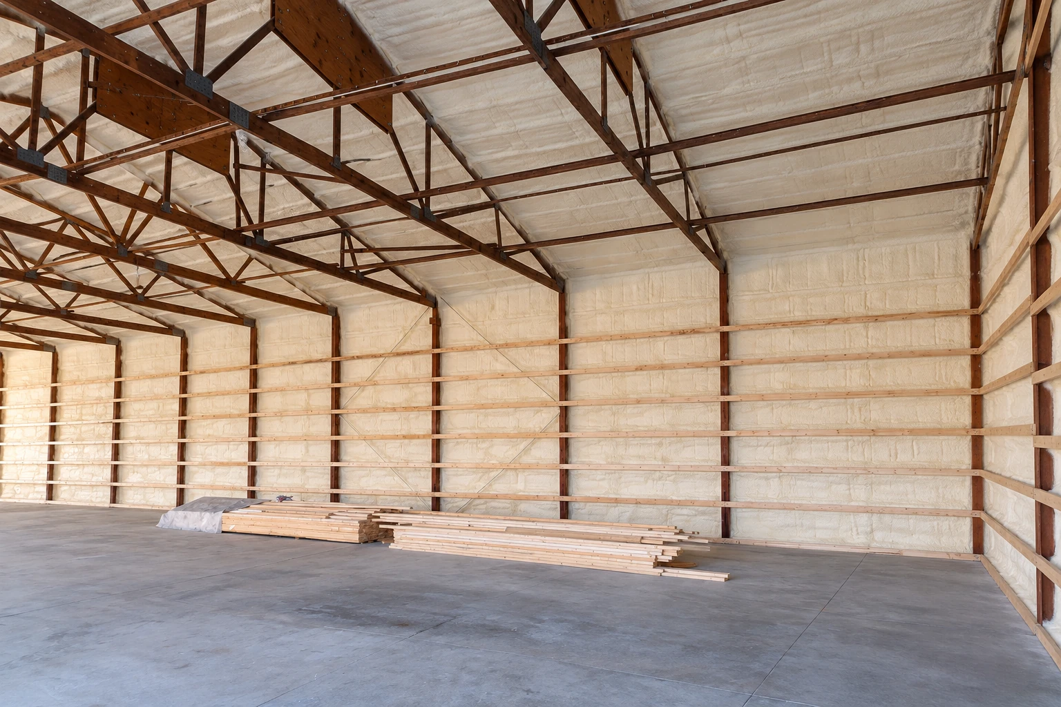 A commercial building interior with white spray foam insulation on the roof and walls, exposed brown trusses, and lumber stacked on the floor.