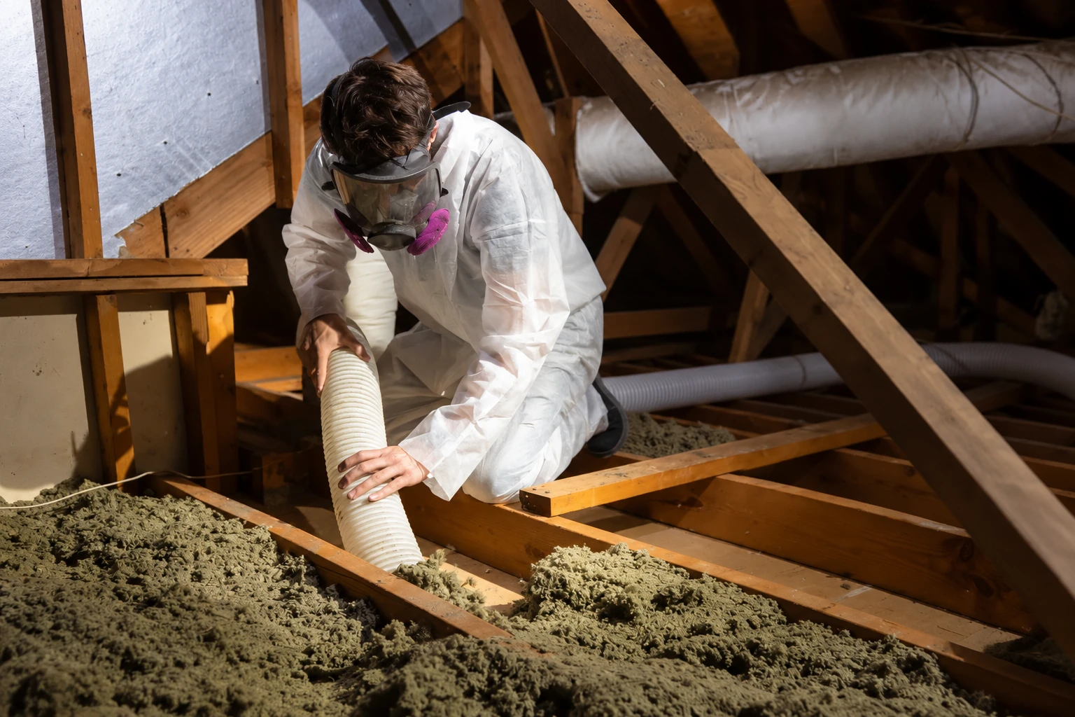 A worker in protective gear vacuums loose attic insulation through a hose between wooden joists.