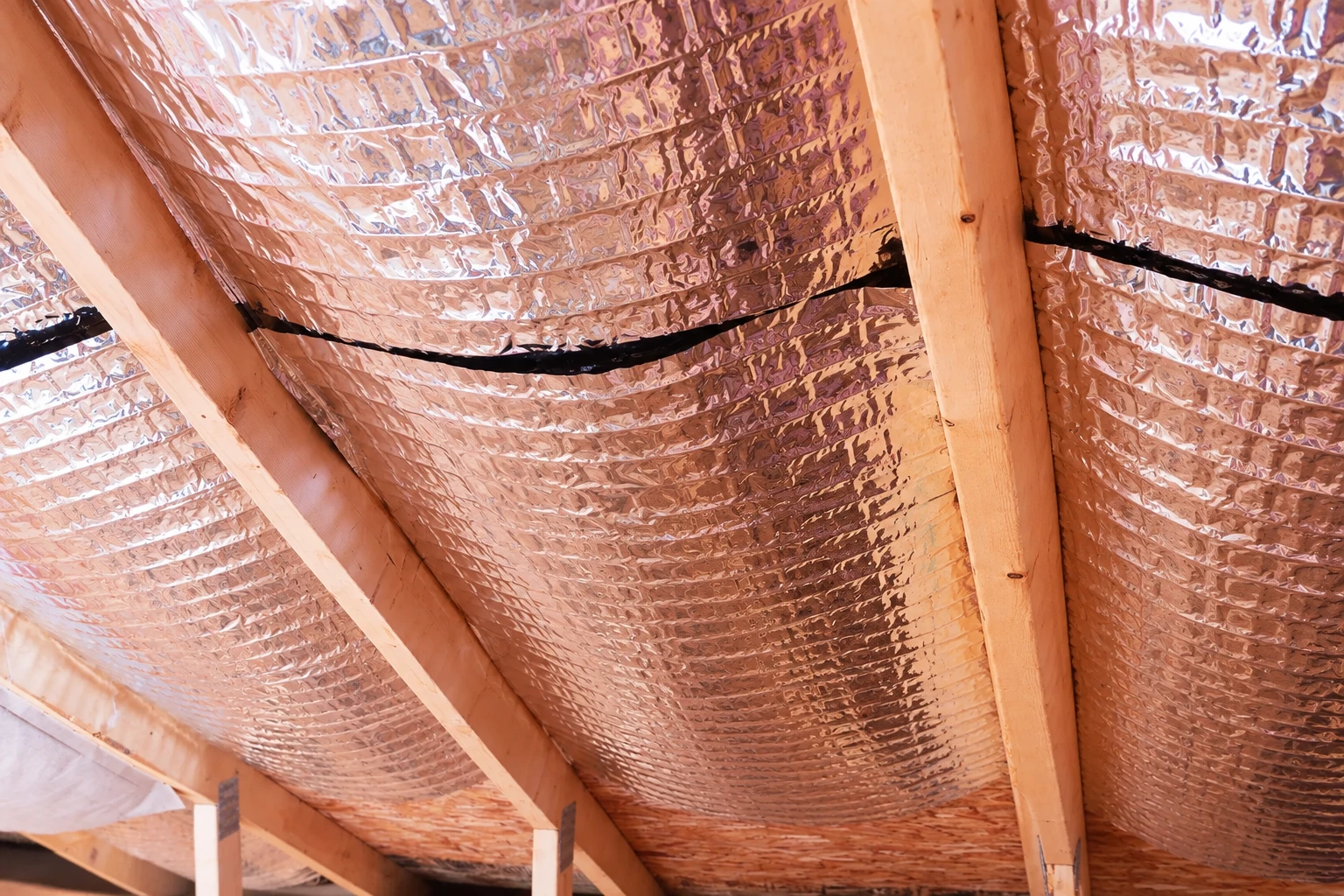 Reflective foil vapor barrier is installed between wooden rafters along a sloped ceiling.
