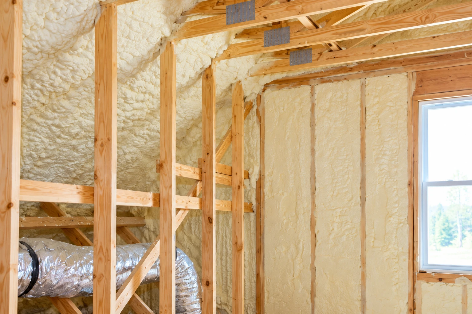 Unfinished framed wall with cream-colored spray foam insulation between studs and a window at right.